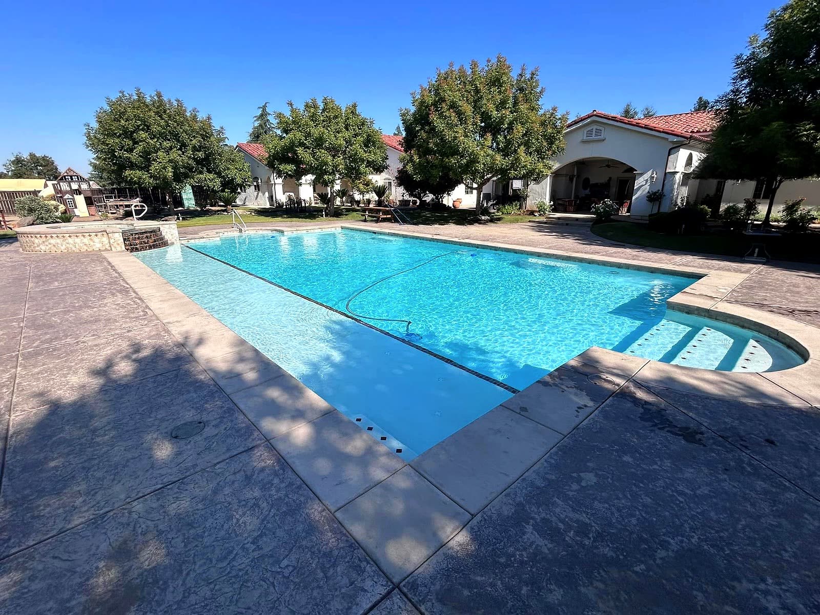Large rectangular pool with attached spa, wide stamped concrete deck and tile-roof home behind