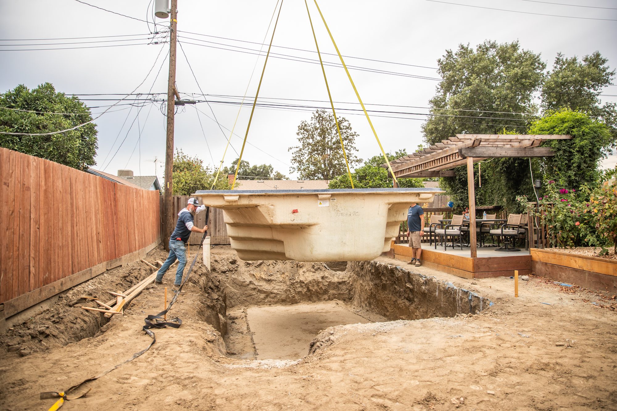 Crane lowering a Latham fiberglass pool shell into an excavated backyard, Neighborhood Pool Service crew guiding it in